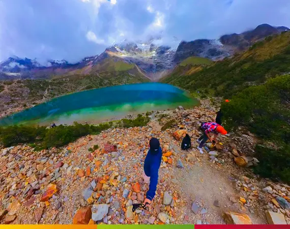 Panoramablick auf die Laguna Humantay in Peru mit türkisfarbenem Wasser, schneebedeckten Bergen und Wolken; steiniger Wanderweg mit Wanderern im Vordergrund.
